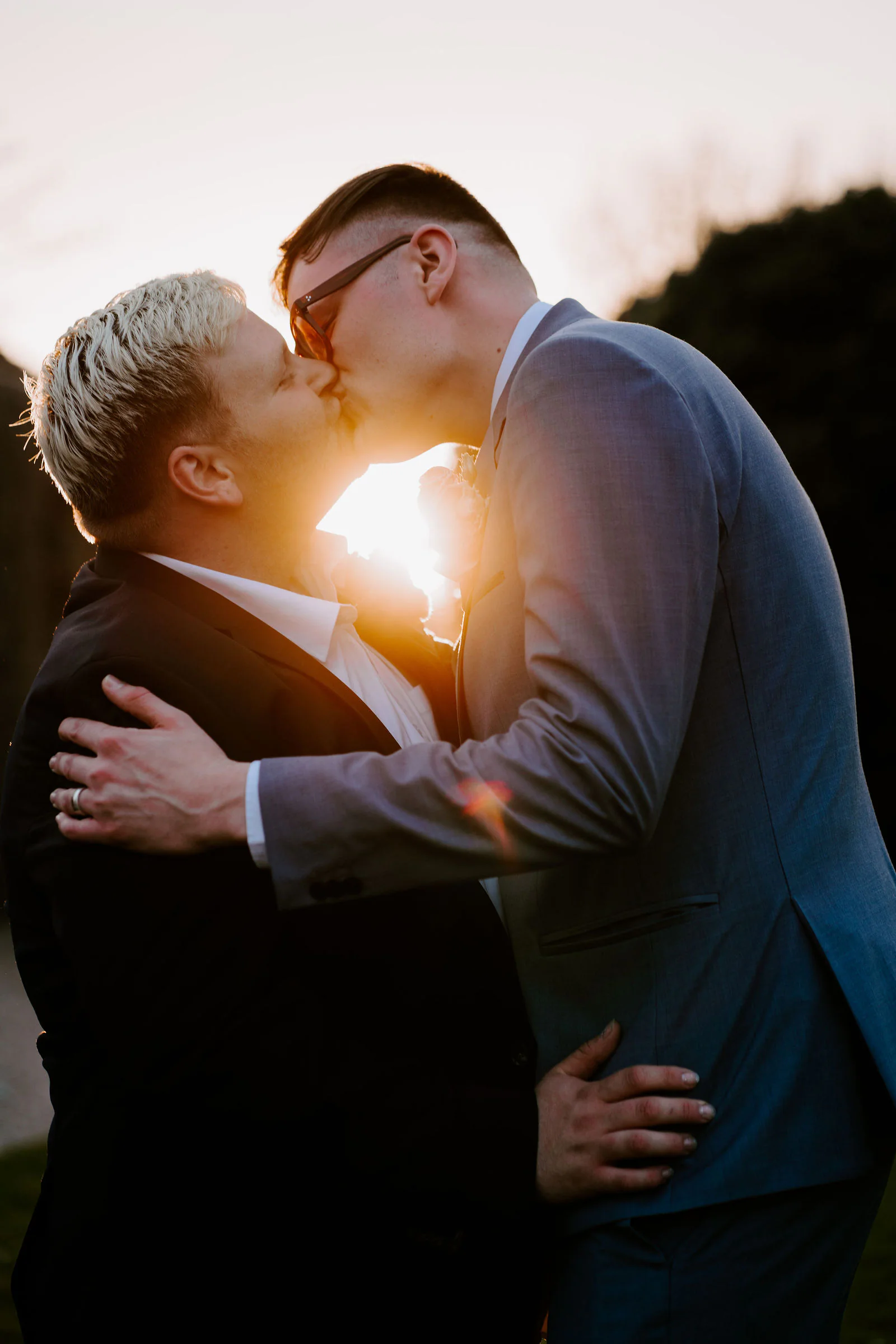 Two grooms kiss in silhouette with low golden sunlight flaring between their faces and dark hedges behind them