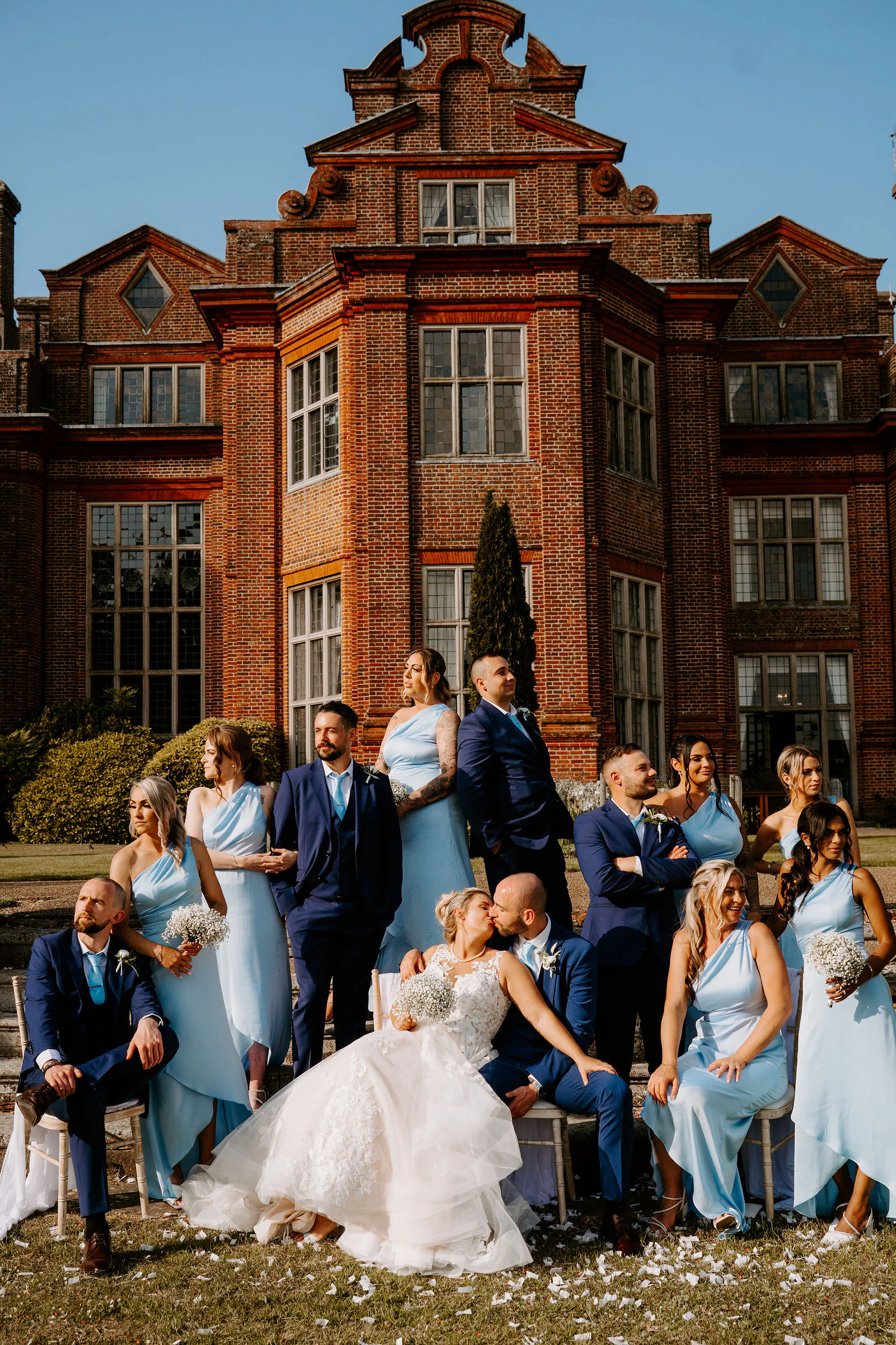 A bride and groom kiss on the lawn in front of the red-brick mansion at Broome Park Hotel while the wedding party sits and laughs around them