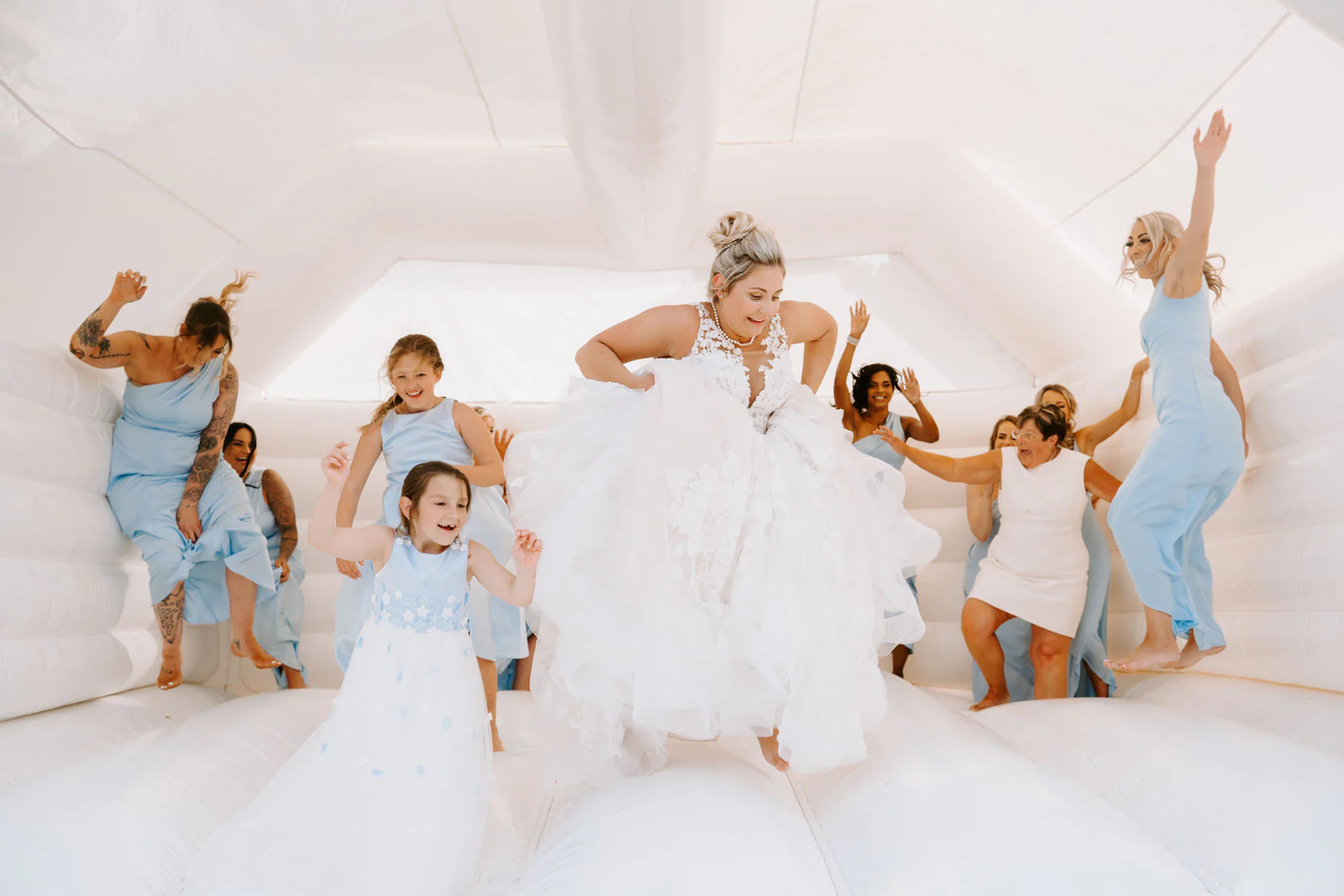 A bride in a full white gown jumps on a white bouncy castle with bridesmaids and children in pale blue at Broome Park Hotel near Canterbury