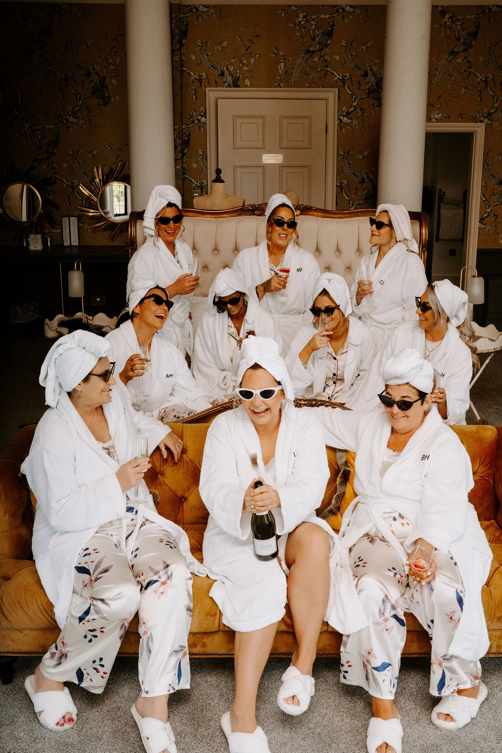 Bridesmaids in white robes and towel turbans pose on a sofa wearing sunglasses at Broome Park Hotel near Canterbury