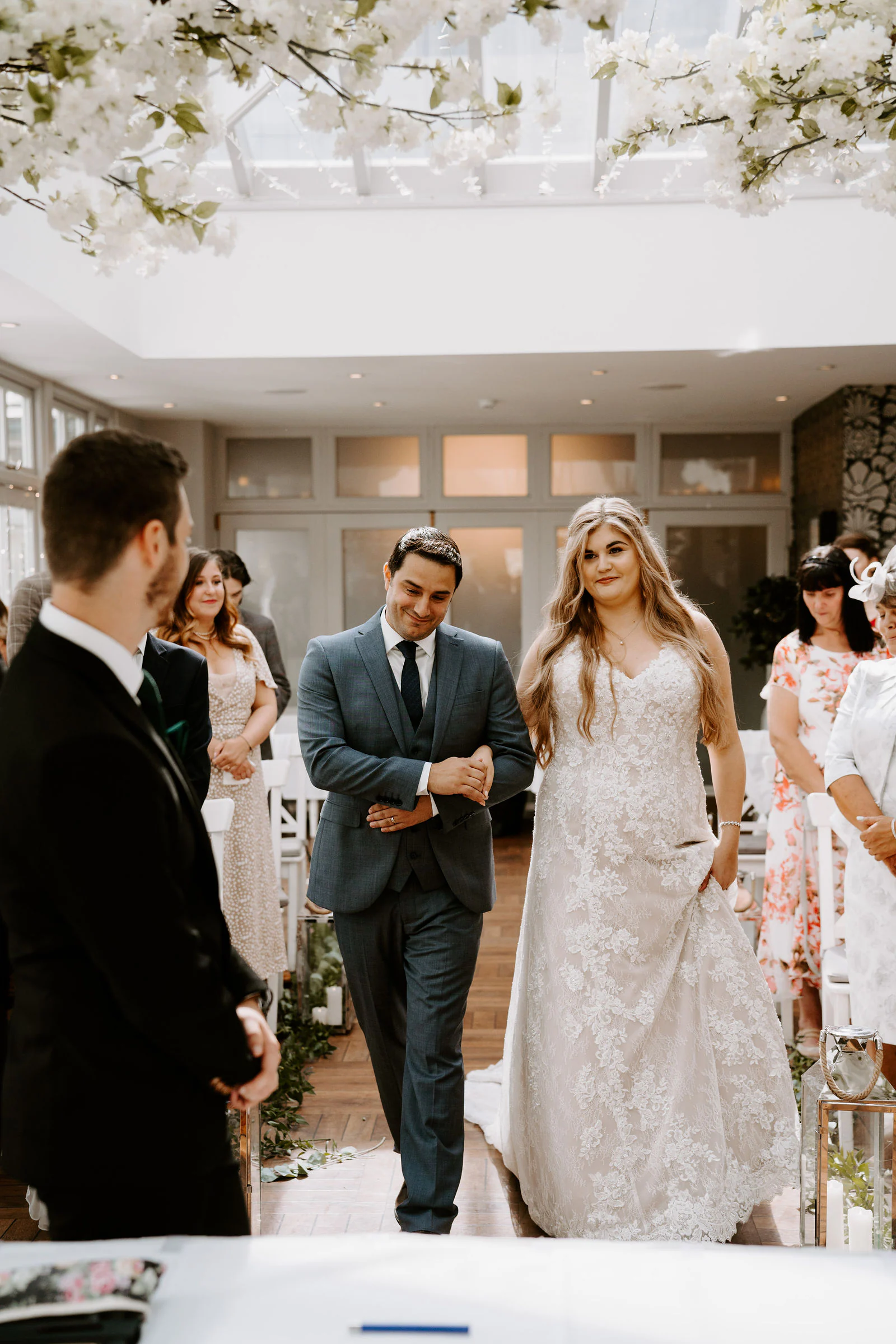 A bride in a lace gown walks down the aisle in a conservatory hung with white flowers in Windermere