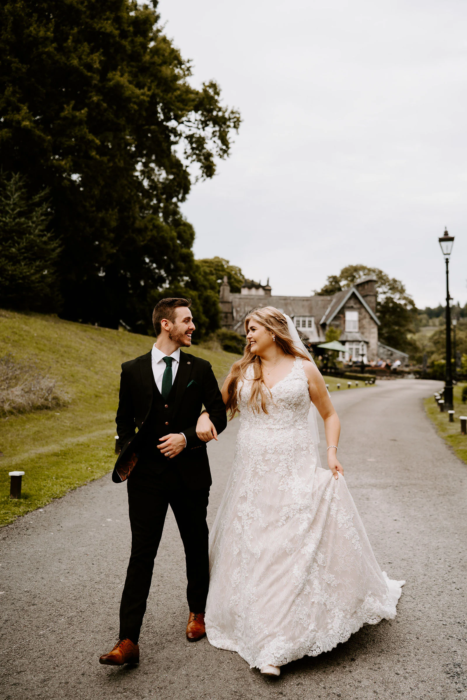 A bride and groom walk arm in arm along a lamplit drive at a country house in Windermere