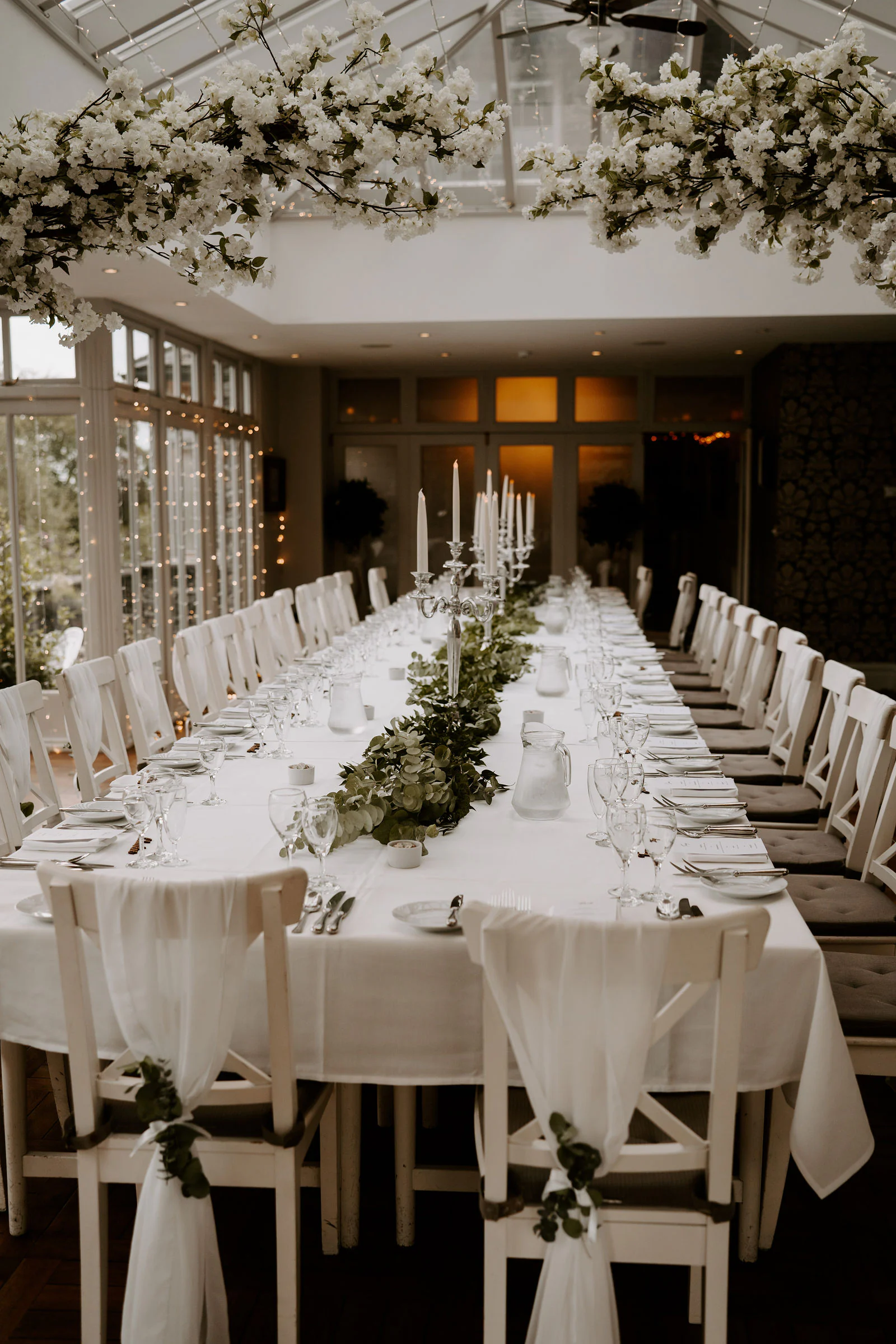 A long wedding table laid under a glass roof with white flowers and linen in Windermere