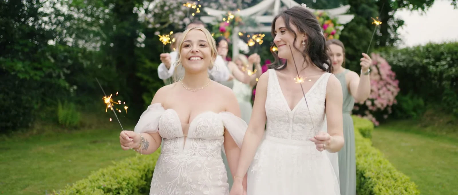 Two brides in lace gowns walk hand in hand between clipped hedges holding lit sparklers, smiling, with bridesmaids in sage green and a white garden pavilion behind them at Cartmel Old Grammar