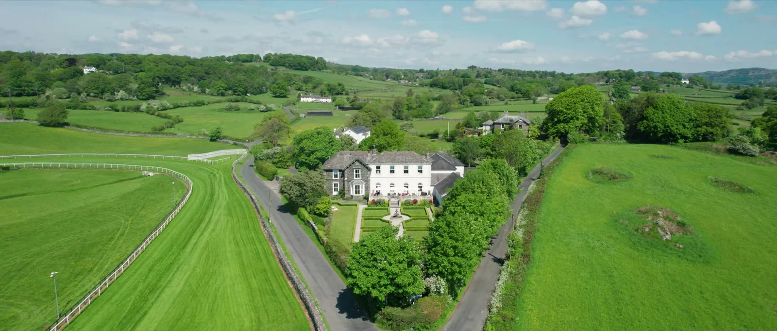 An aerial view of a white Georgian house with a formal garden of clipped hedges and a fountain, beside a racecourse and green fields with rolling hills beyond at Cartmel Old Grammar