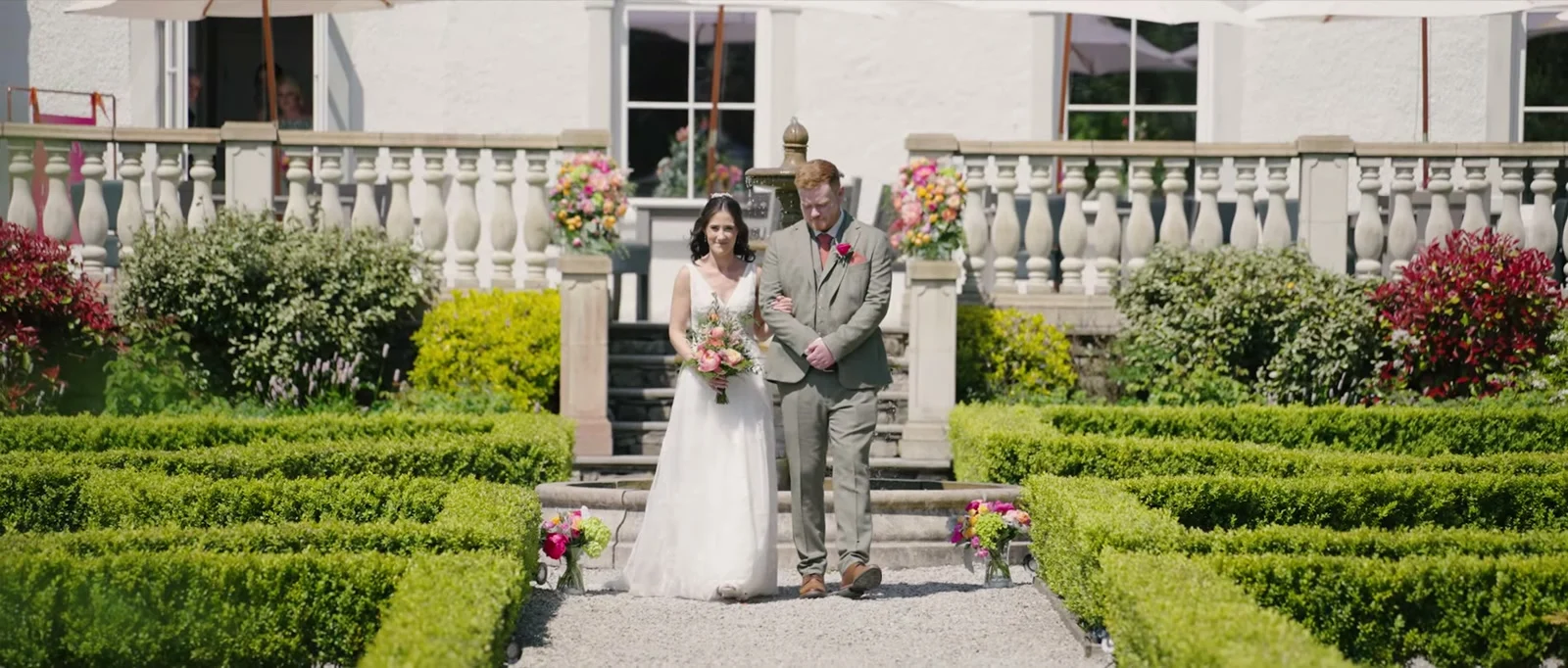 A bride in a white dress carrying a pink and peach bouquet walks arm in arm with a man in a grey-green suit along a gravel path between clipped box hedges, with a stone balustrade and fountain behind at Cartmel Old Grammar
