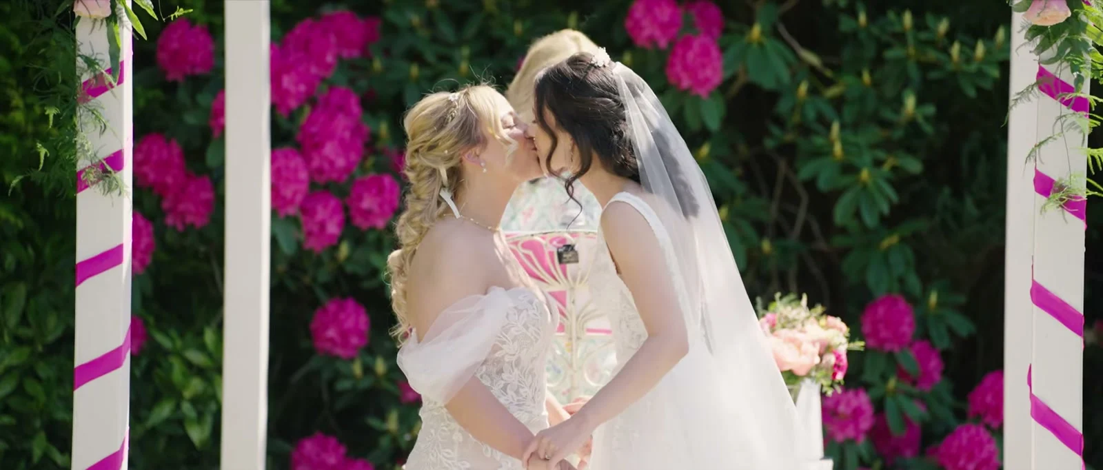 Two brides kiss under a white pavilion wound with magenta ribbon, in front of deep pink rhododendrons, one in a veil and one in an off-the-shoulder lace gown at Cartmel Old Grammar