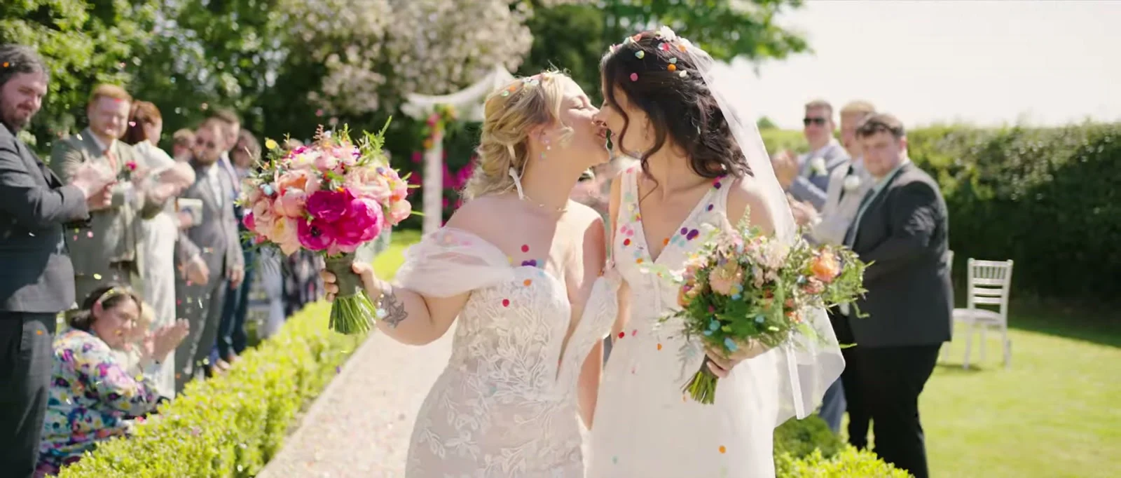 Two brides kiss on a gravel path as colourful confetti falls, each holding a bouquet, with clapping guests beside a clipped hedge and a white pavilion behind at Cartmel Old Grammar