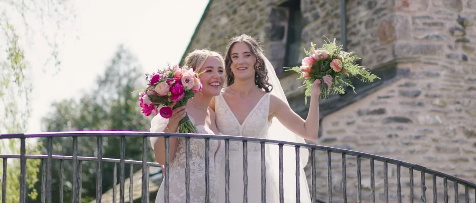 Two brides in lace gowns, each holding a pink bouquet, lean together smiling at a curved iron railing in front of a stone building at Cartmel Old Grammar