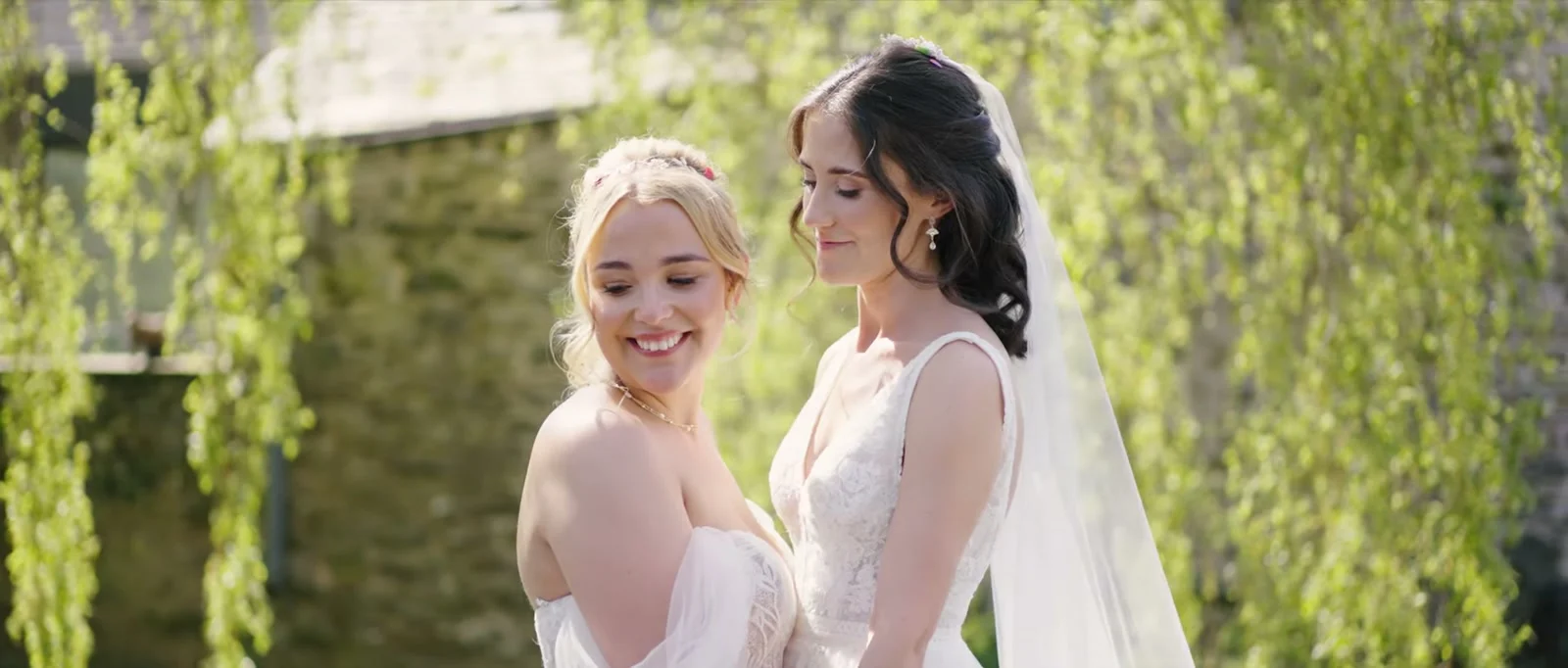 Two brides stand close together among hanging green willow branches in sunlight, one in an off-the-shoulder lace gown smiling downwards and the other in a veil looking down at Cartmel Old Grammar
