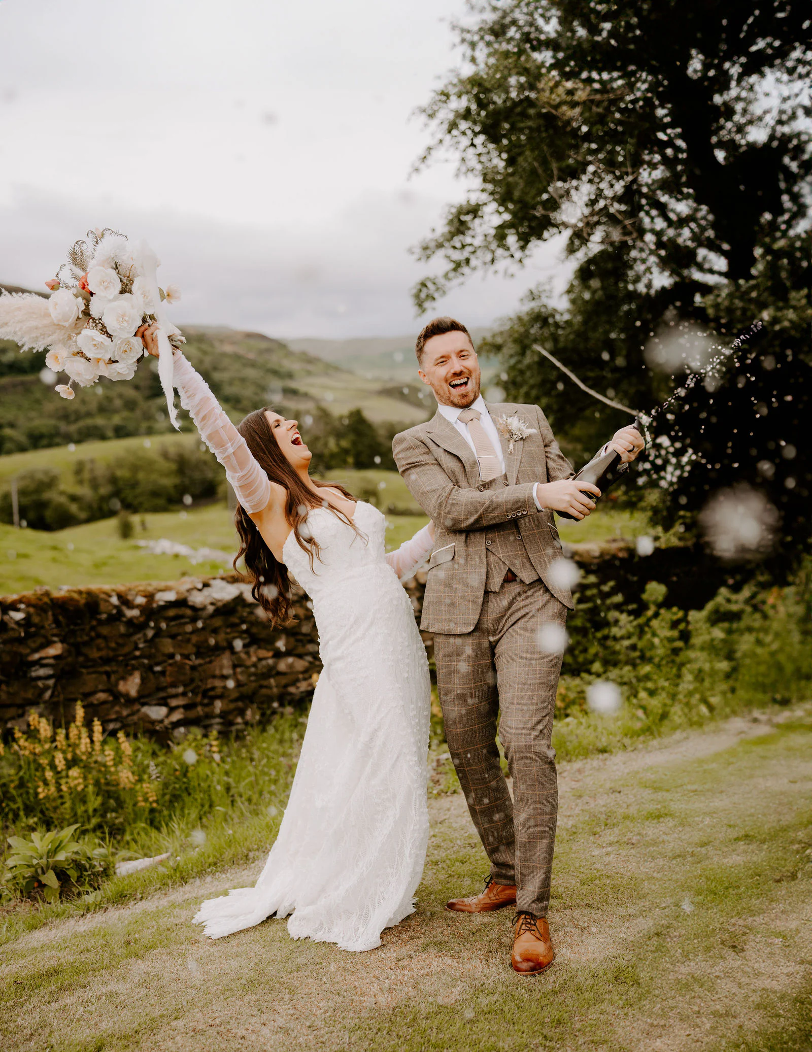 A bride raises her bouquet and a groom raises his arms in celebration in a field with hills behind at Fairbank Barn in Staveley