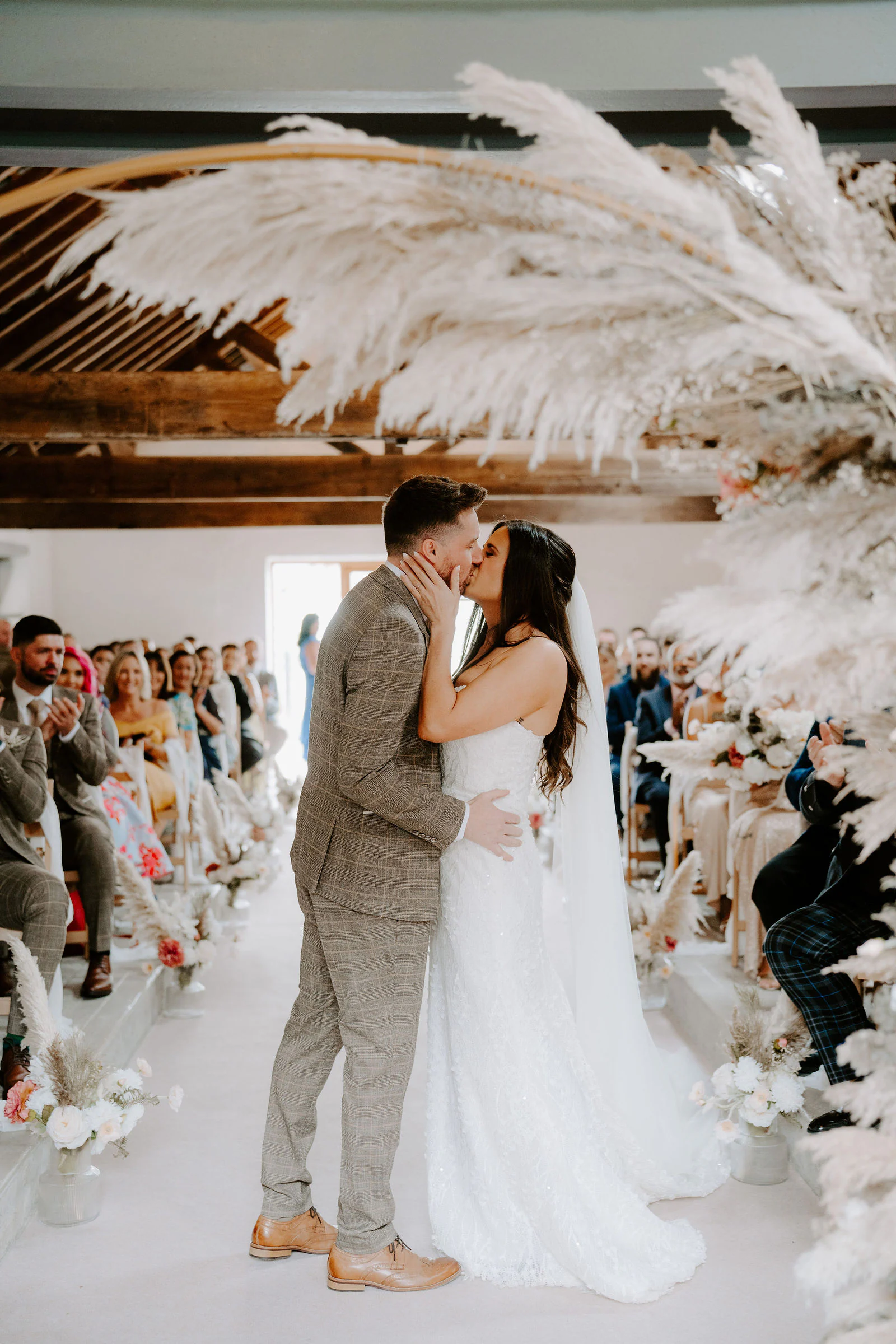 A bride and groom kiss at the end of the aisle beneath a pampas grass arch at Fairbank Barn in Staveley