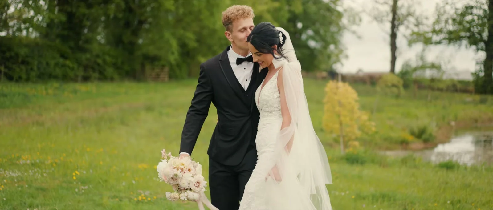 A groom in a black dinner suit and bow tie kisses the top of his bride's head as they walk through a meadow beside a pond, her long veil lifting behind her and a bouquet of cream and blush flowers in his hand at Holesfoot