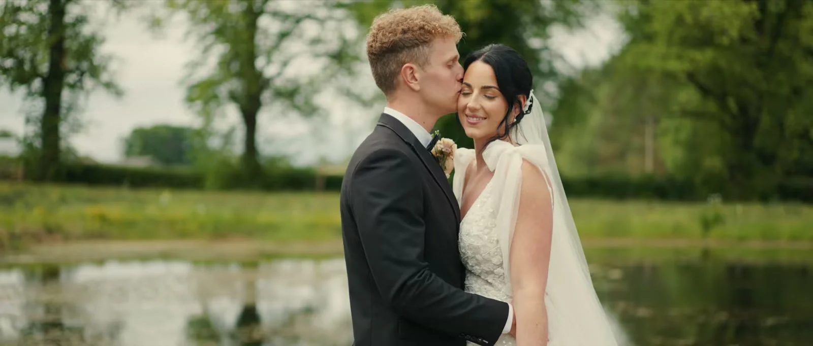A groom rests his lips against his bride's forehead as they embrace beside a pond with trees blurred behind them at Holesfoot