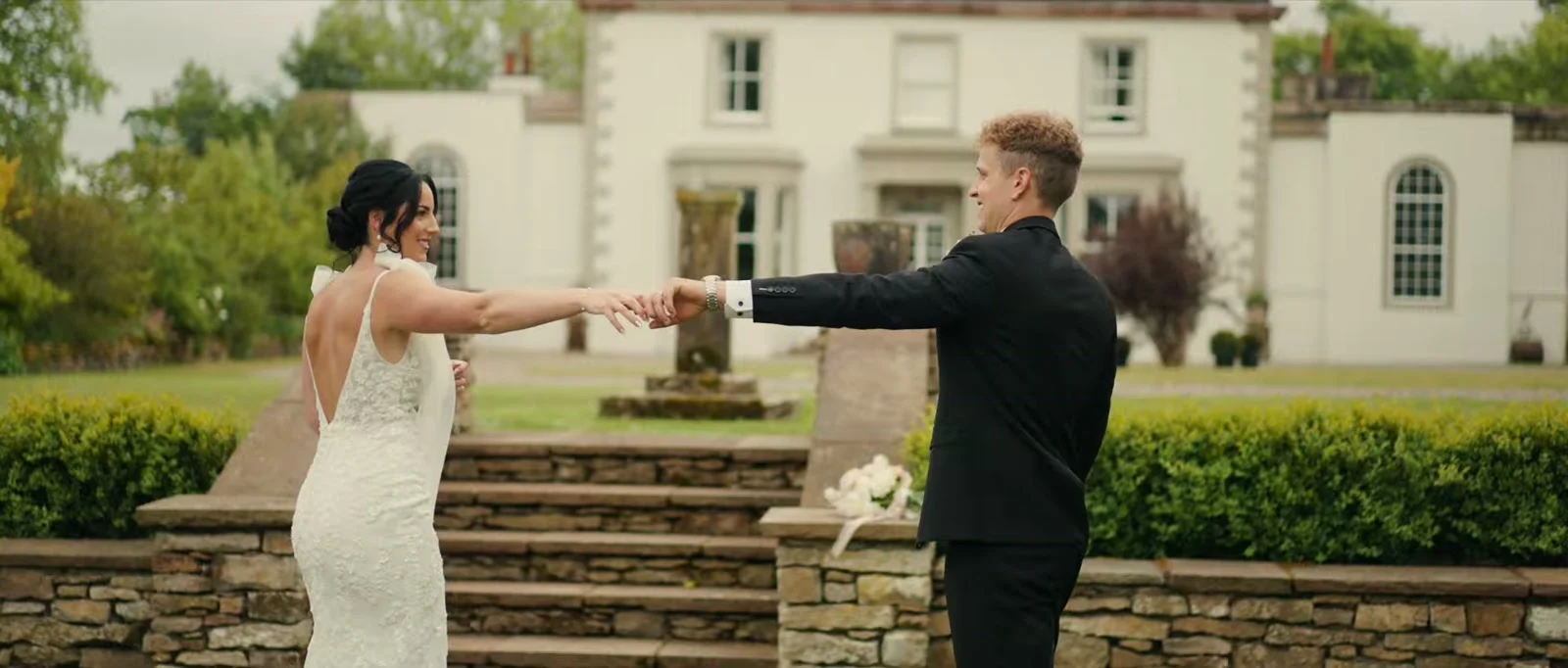 A bride in a lace gown with an open back turns as her groom in a black suit takes her hand on stone steps, with clipped hedges and the pale manor house behind at Holesfoot