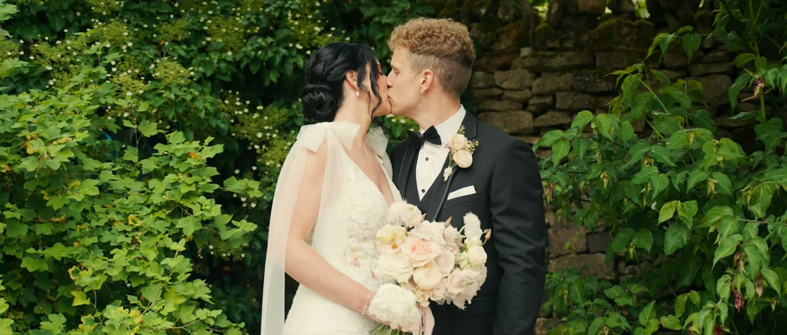 A bride and groom kiss against green leaves and an old stone wall, she holding a bouquet of cream and peach roses with soft tulle tied at her shoulder at Holesfoot