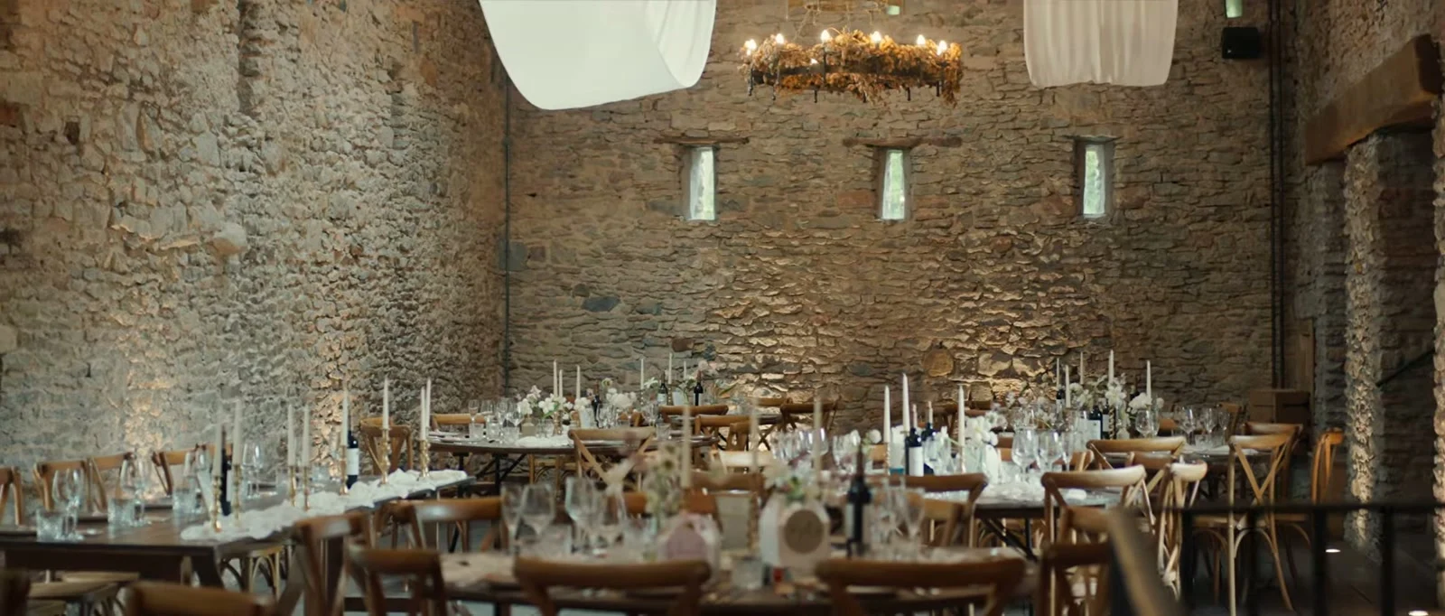 A stone barn at Holesfoot set for a wedding breakfast with long tables, tall candles, cross-back chairs, a candle chandelier and white fabric hanging from the roof beams