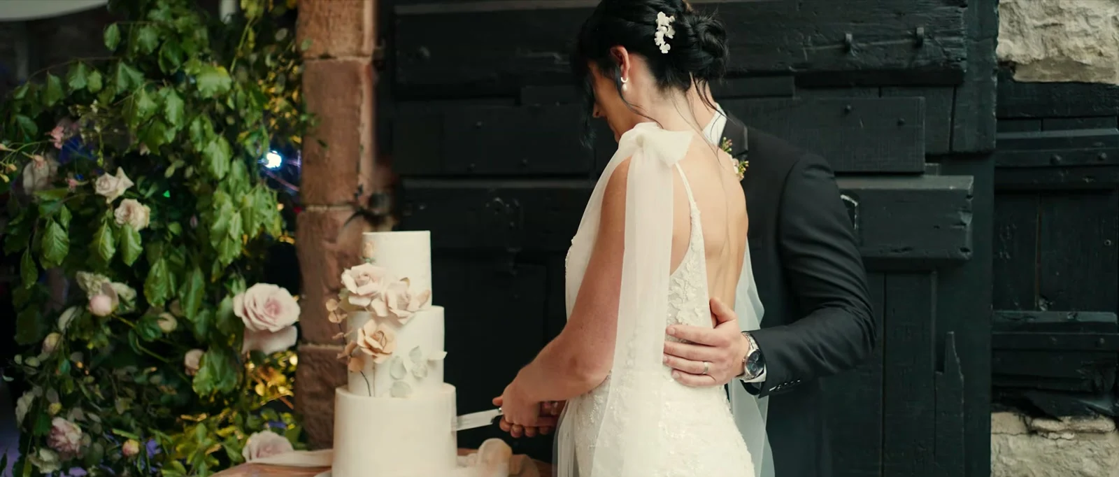 A bride and groom cut a tall white wedding cake decorated with sugar flowers in front of a black timber door and a climbing plant with pale roses at Holesfoot