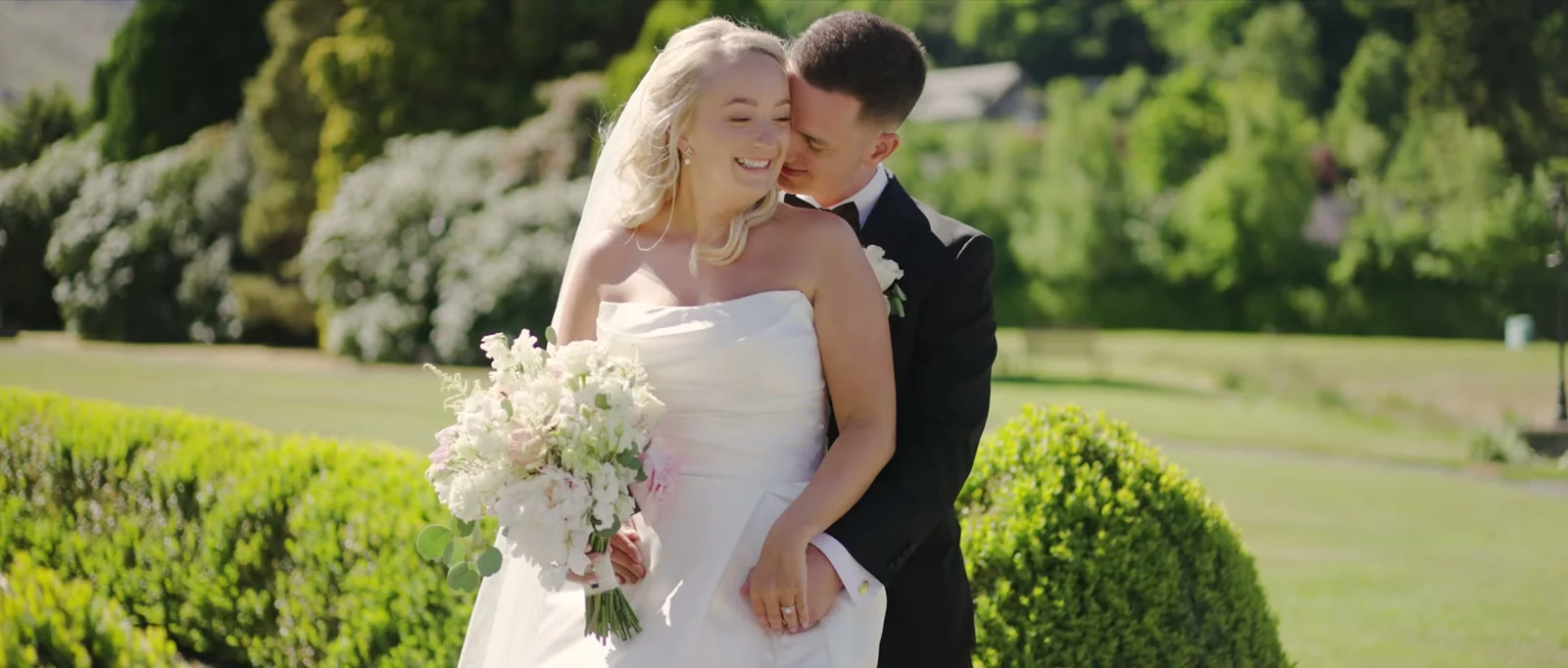 A smiling bride in a veil holding a bouquet of white and blush flowers is held from behind by her groom in a black suit in a sunlit garden with clipped hedges at Inn On The Lake