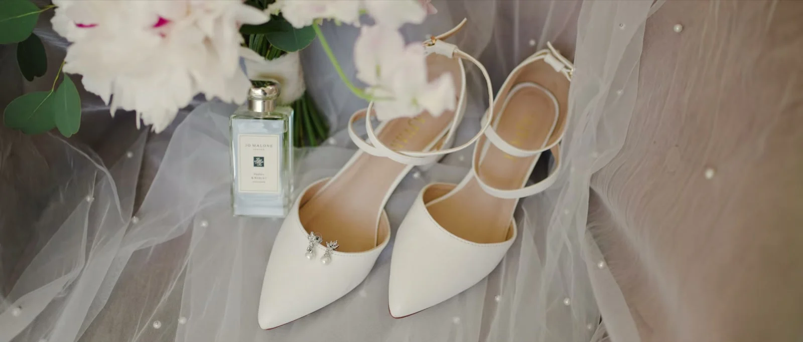 White pointed ankle-strap shoes with pearl drop earrings on one toe, a bottle of perfume and a bouquet of white and blush flowers resting on a veil dotted with pearls