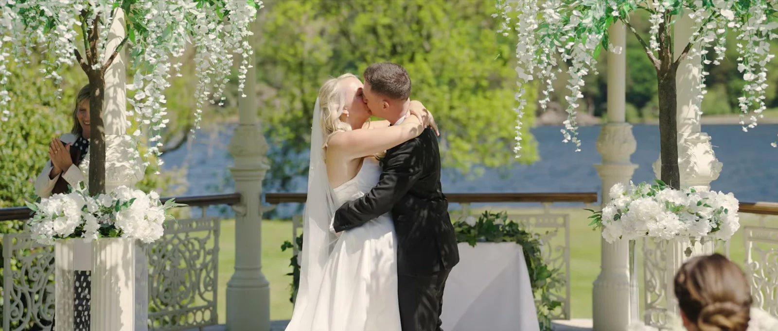 A bride in a veil and a groom in a black suit kiss in an embrace inside a gazebo hung with white blossom, with the lake and a lawn behind the pillars