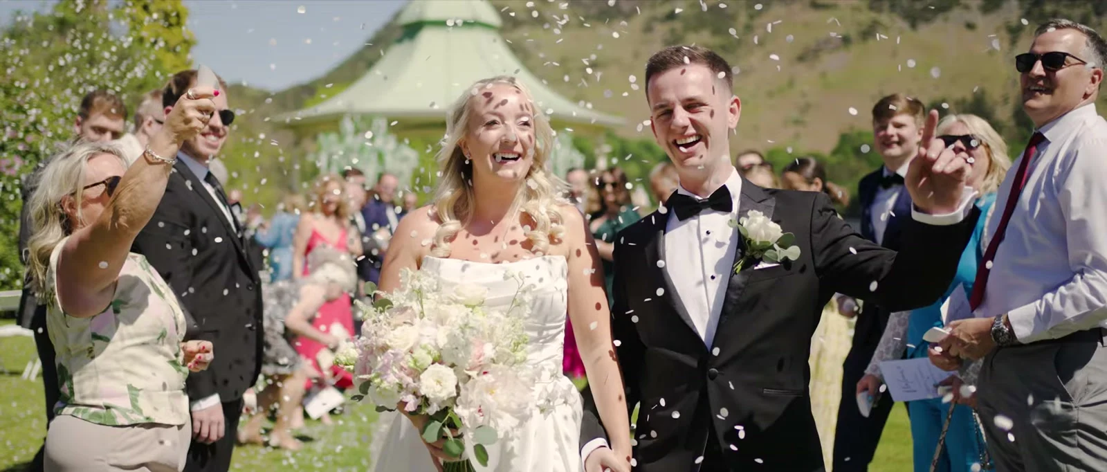 A laughing bride holding a bouquet of white and blush flowers walks beside her smiling groom as guests throw confetti, with a green-roofed gazebo and a fellside behind them
