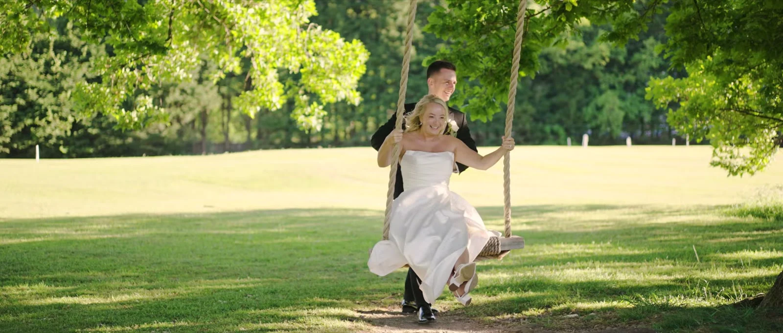 A groom in a black suit pushes his smiling bride on a rope swing hung from a tree over a sunlit lawn, her white dress lifting as she swings