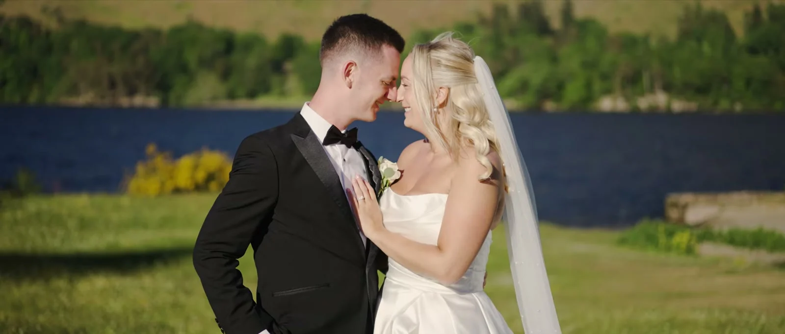 A groom and a bride in a long veil touch noses and smile in front of the dark blue water of Ullswater with green slopes behind