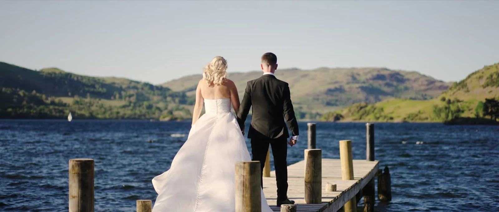 A bride in a long white dress and a groom in a black suit walk hand in hand along a wooden jetty over blue lake water towards the hills