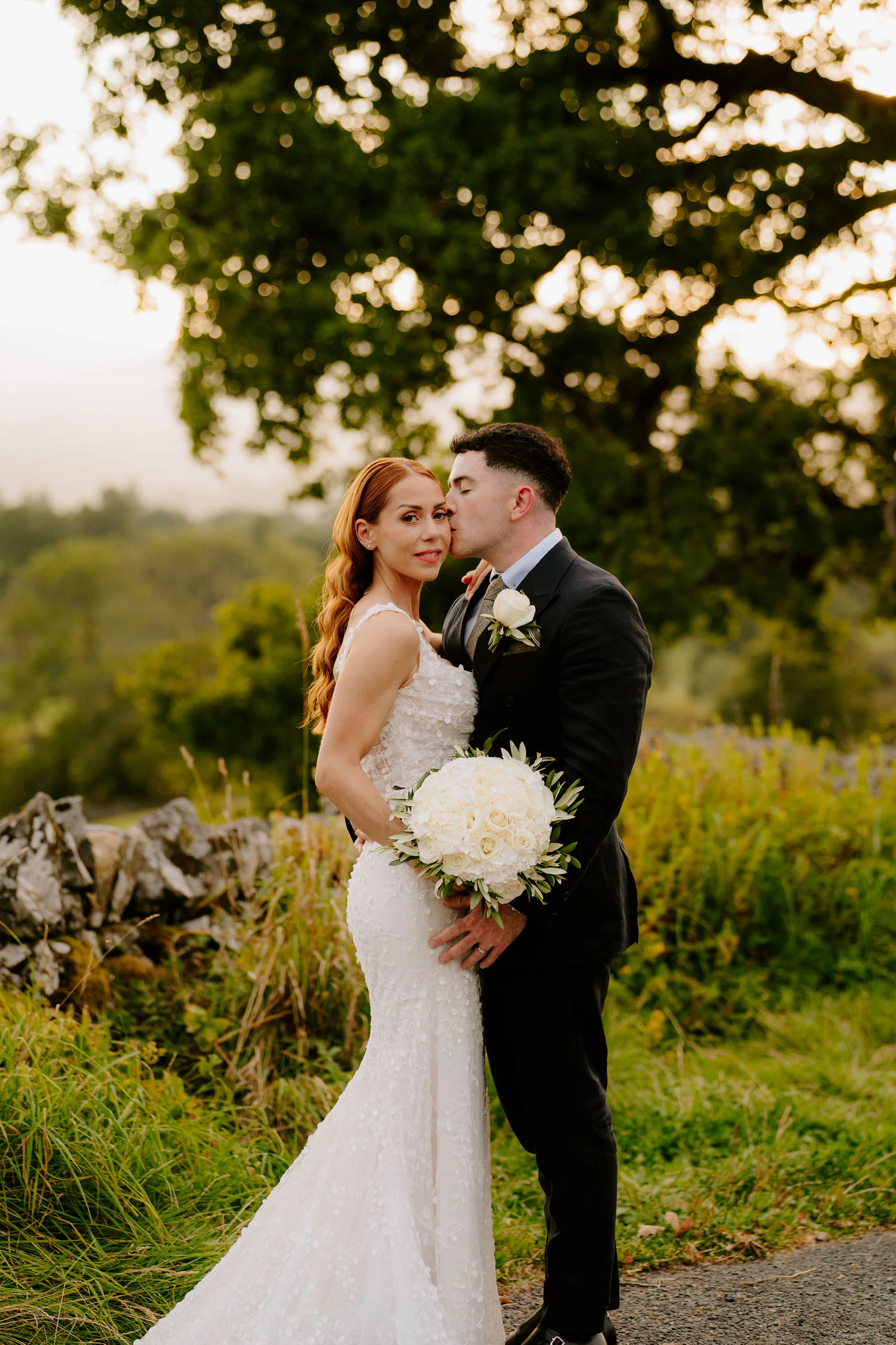 A bride and groom kiss in warm evening light among long grass and trees at Broadoaks near Windermere