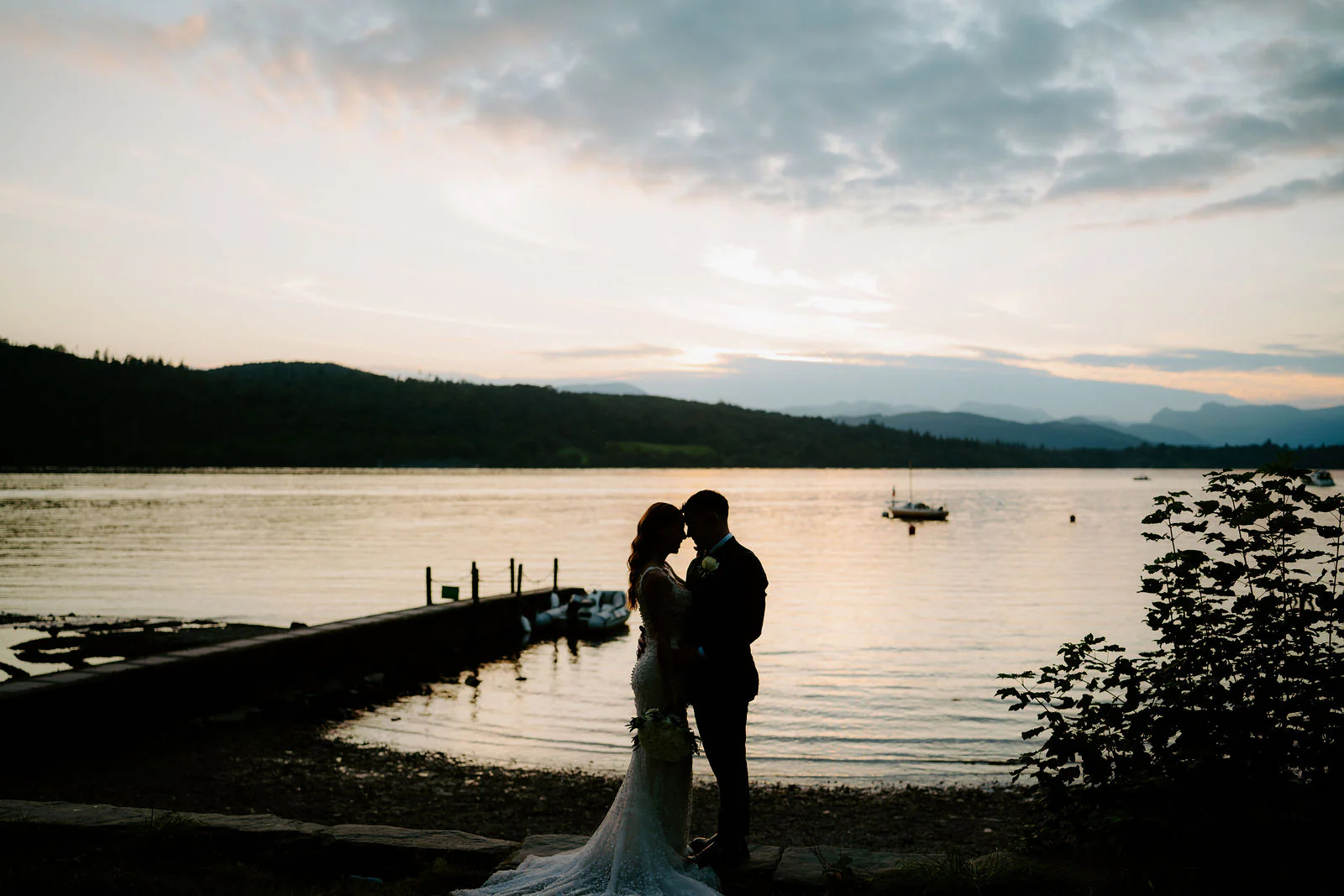 A bride and groom embrace in silhouette on a jetty beside Windermere at sunset