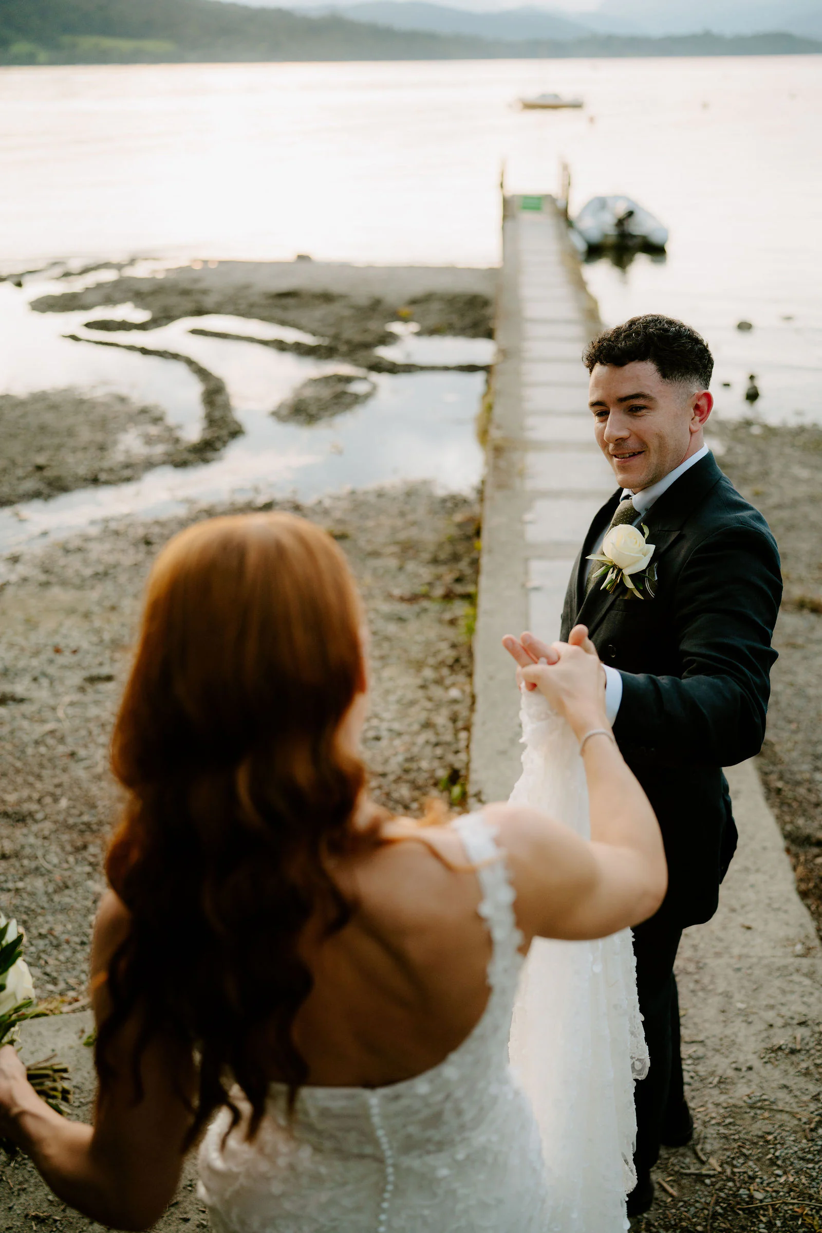 A bride leads her groom by the hand along a wooden jetty beside the lake at Broadoaks near Windermere