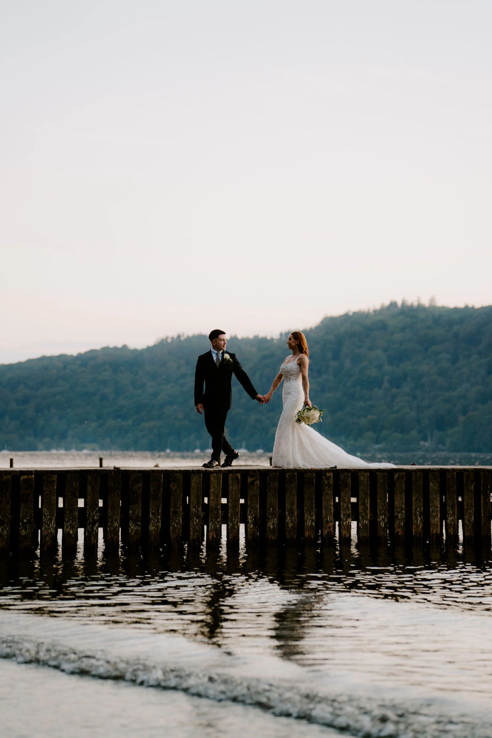 A bride and groom walk along a jetty above the shoreline with wooded hills across the water near Windermere