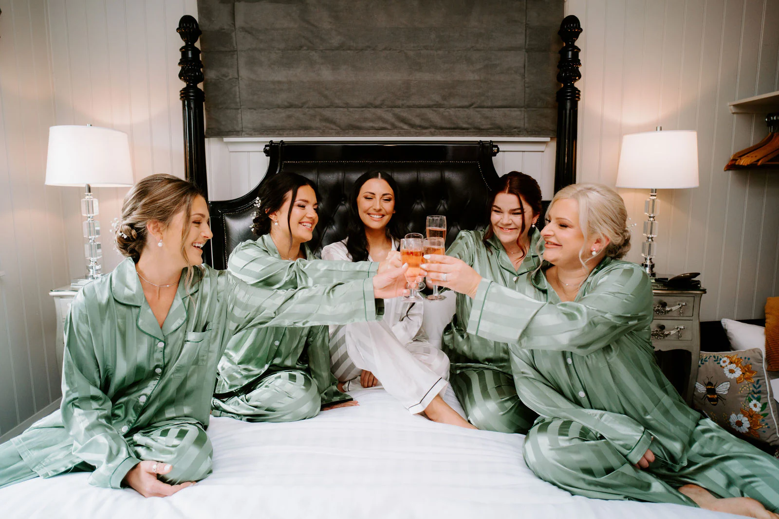 Bridesmaids in green satin robes raise glasses together on a bed in the bridal suite at Broadoaks Country House