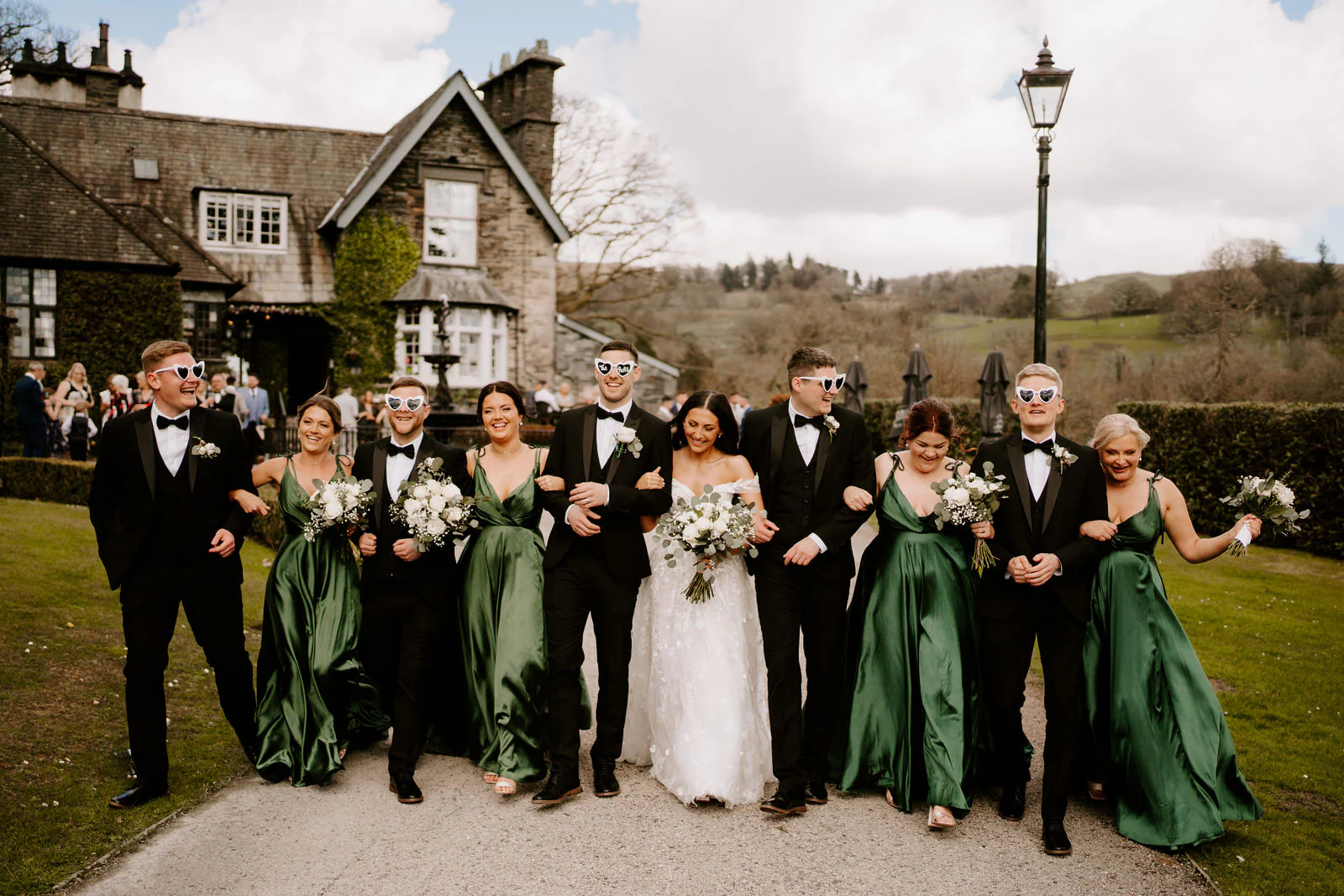 The wedding party stand in a line in front of the stone house at Broadoaks Country House, the bridesmaids in green dresses