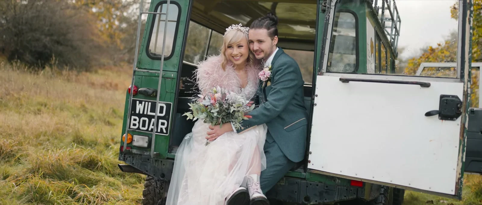 A smiling bride in a sparkling headpiece and a pink feathered wrap sits close to her groom on the back of an open green Land Rover with a WILD BOAR number plate, a bouquet of pastel flowers in her lap, in golden autumn grass