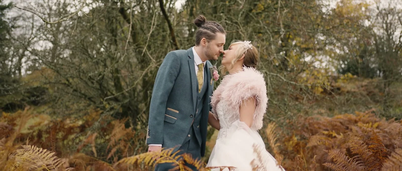 A groom in a blue-grey suit and a bride in a pink feathered wrap kiss among golden bracken and bare autumn trees near The Wild Boar