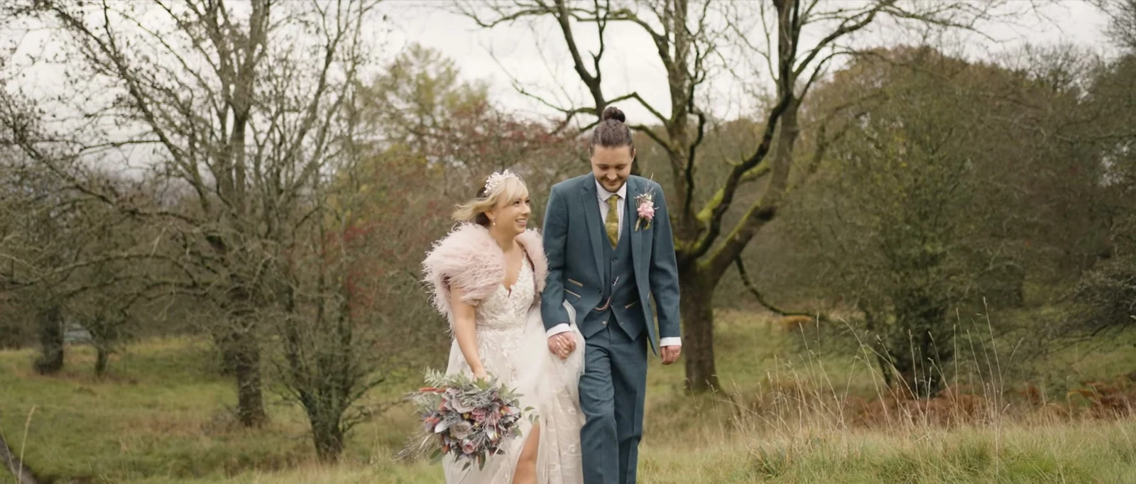 A bride carrying a bouquet and a groom with a flower on his jacket walk side by side across open parkland with bare trees near The Wild Boar