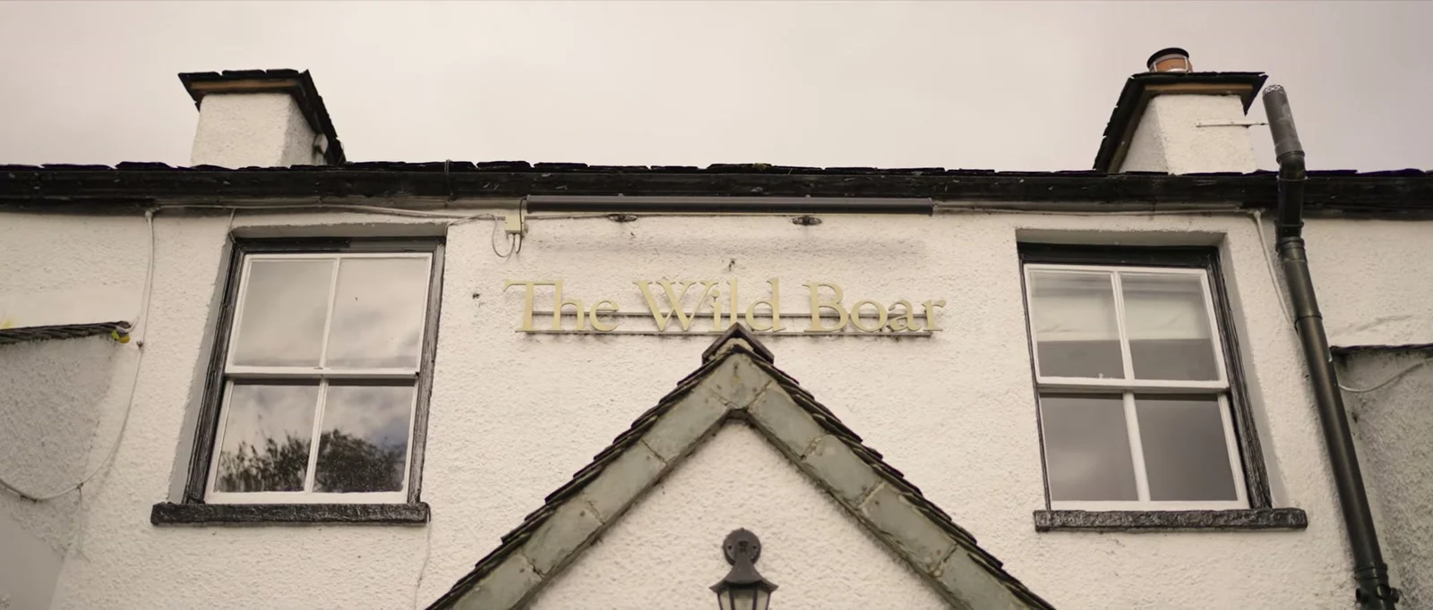 The white front of The Wild Boar with gold lettering above a gable window and two chimneys under a grey sky