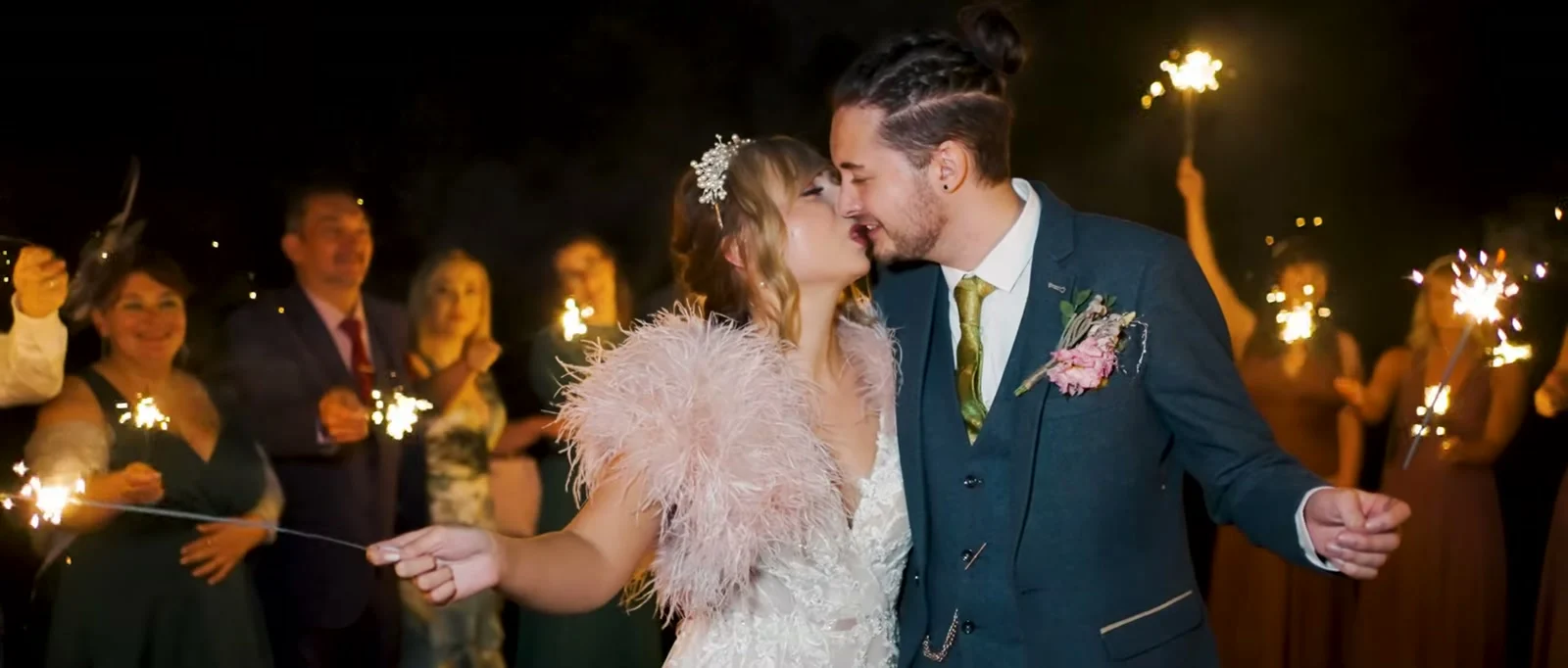 A bride in a pink feathered wrap and a groom in a blue suit kiss at night holding sparklers, with smiling guests holding sparklers around them