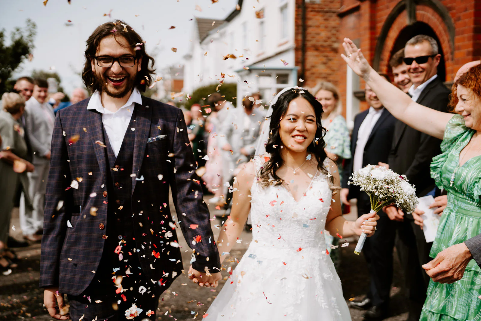 Anthony in a checked suit and glasses and Jasmine in a long veil walk through confetti thrown by their guests in front of a red-brick building with white bay windows