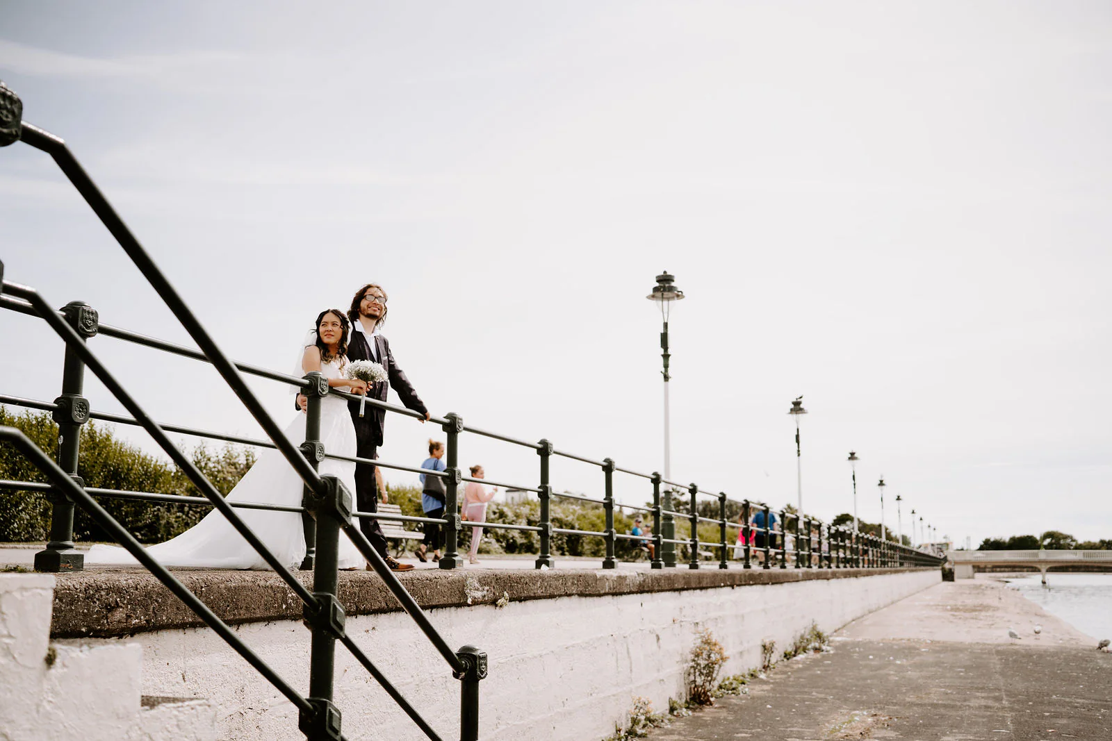 Anthony and Jasmine stand small beside black railings on a long seafront promenade with lamp posts and a wide cloudy sky