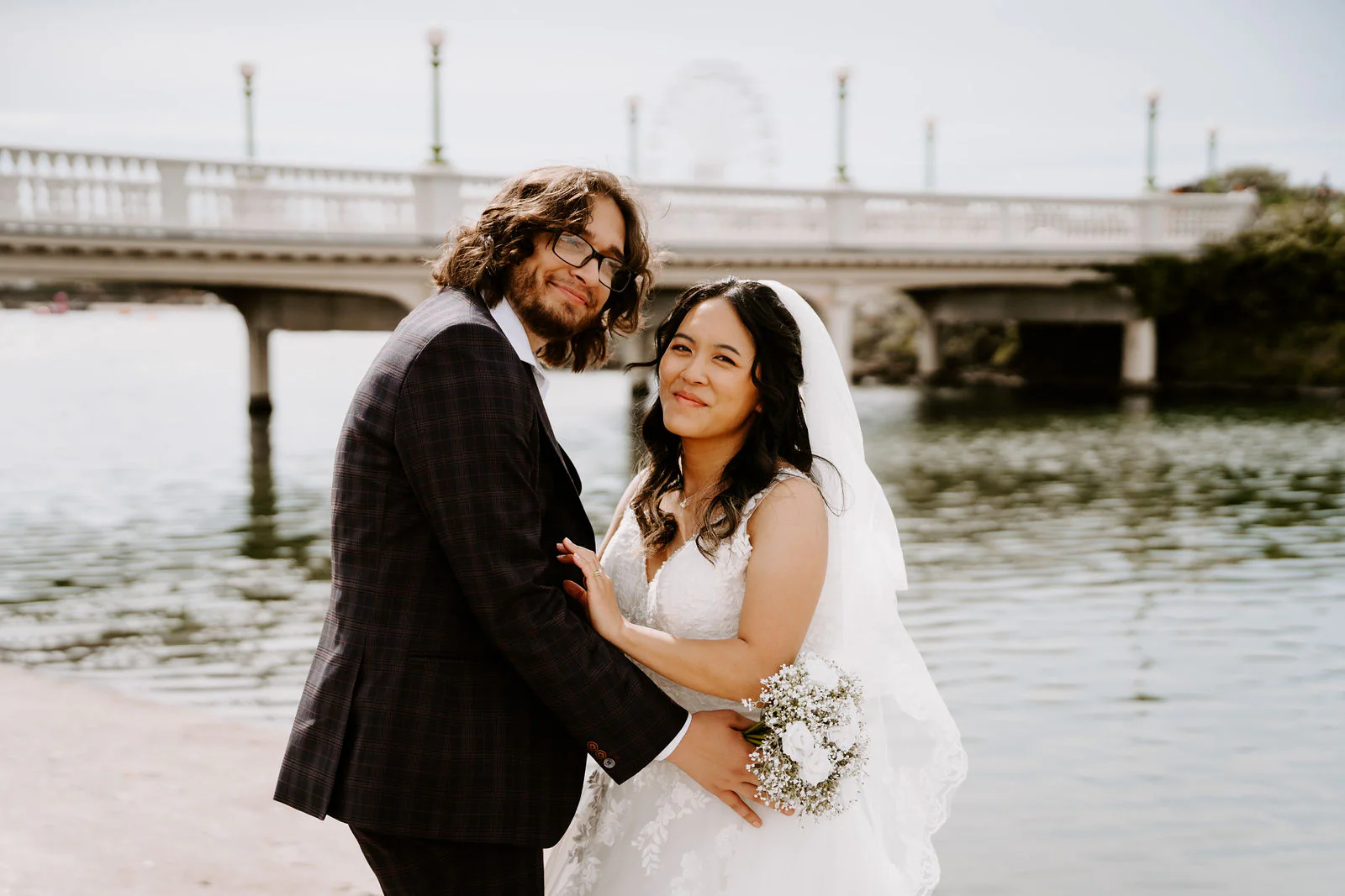Anthony and Jasmine hold each other beside the water with a balustraded bridge behind them, Jasmine holding a small bouquet