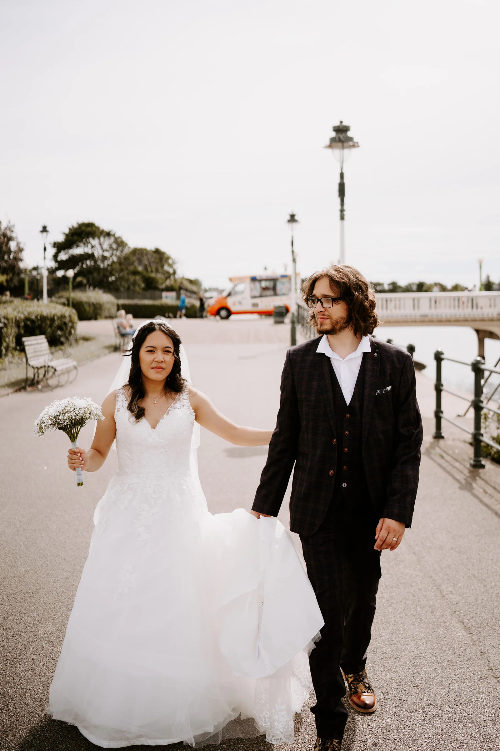 Anthony and Jasmine walk along a promenade towards the camera, Jasmine lifting her skirt and holding a small bouquet, with lamp posts behind them