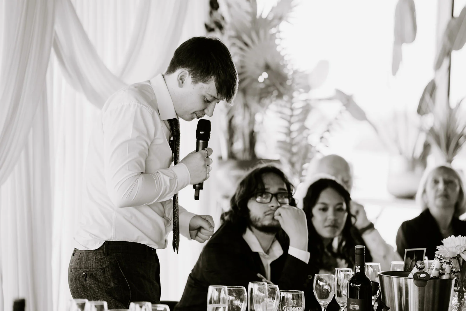 In black and white, a man speaks into a microphone while Anthony and Jasmine listen at the top table, Jasmine wiping a tear