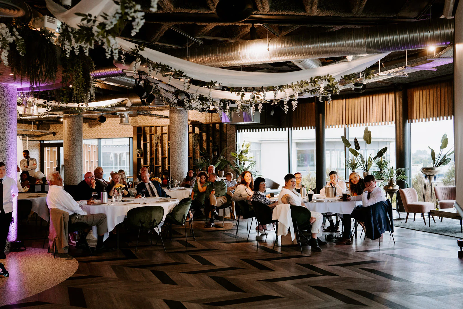 A wide room with garlands of greenery and white fabric hanging above guests at round tables, large windows along one side