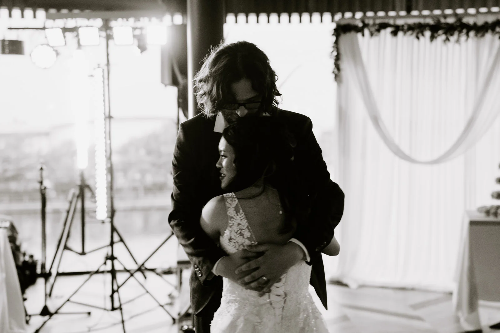 In black and white, Anthony and Jasmine embrace in front of white drapes, with a camera tripod behind them