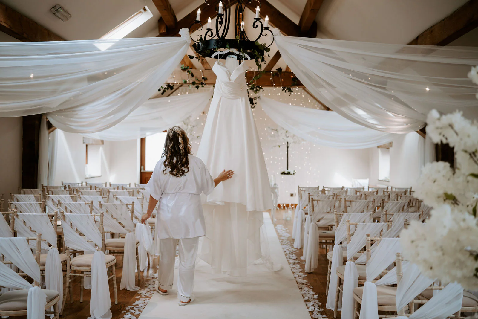 Emma walking into the ceremony room at Beeston Manor, hung with white drapes and lined with chairs, a wedding dress and a chandelier at the far end