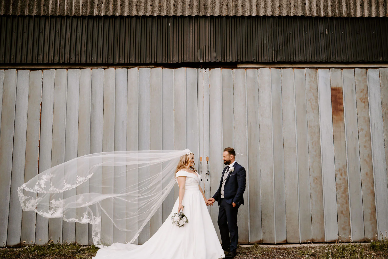 Emma and Sean with her veil catching the breeze in front of a weathered timber wall outside Beeston Manor