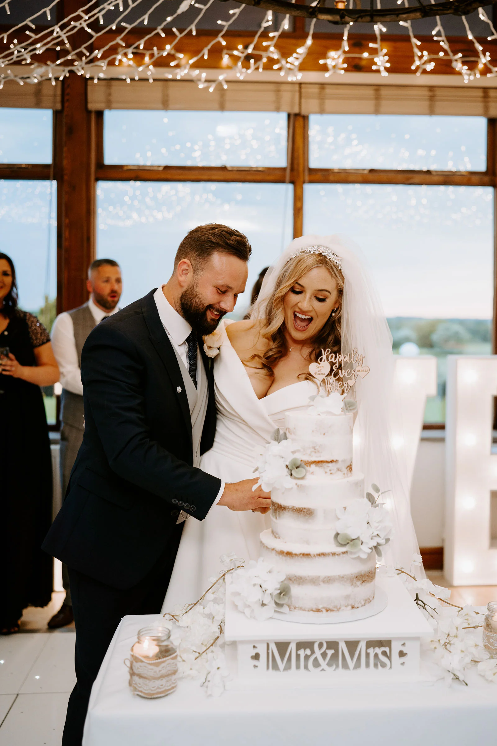 Emma and Sean cutting their wedding cake under fairy lights at Beeston Manor, with a window and tall letters behind them